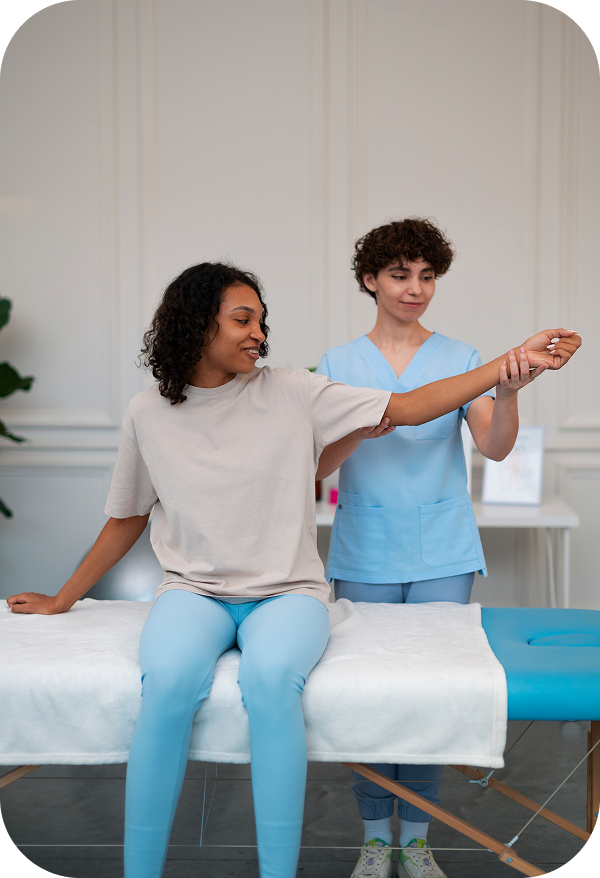 A physiotherapist is sitting on an examination bed with a female patient, showing her an arm exercise to improve her range of motion.